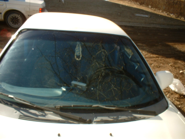 View of a car's windshield and interior, with a sunshade and a dangling air freshener. Another white car is visible to the left