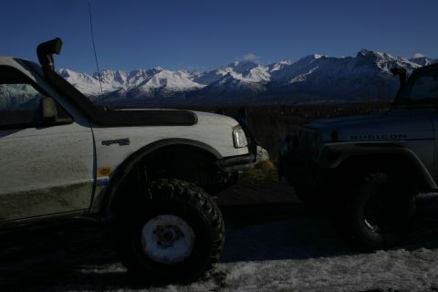 2003 Jeep TJ - Two off-road vehicles, a Jeep Wrangler Rubicon and a white truck with a snorkel, are parked with a backdrop of snow-capped mountains.