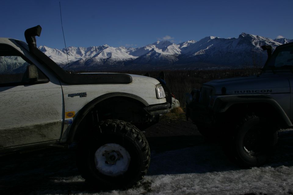 Silver Jeep Wrangler Rubicon with snorkel and white off-road truck parked with snowy mountains behind