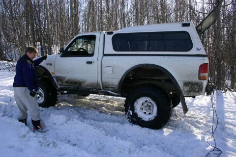 2003 Jeep TJ - A white pickup truck with a camper shell is stuck in deep snow, with a person standing beside it.