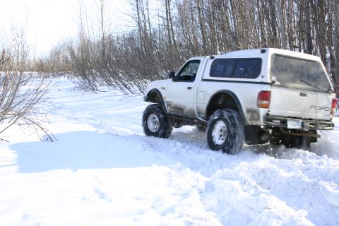 2003 Jeep TJ - White Ford pickup truck with a camper shell navigates a snowy trail, kicking up snow as it drives.