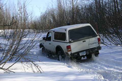 2003 Jeep TJ - White Ford pickup truck with camper shell navigates snowy terrain, kicking up snow.
