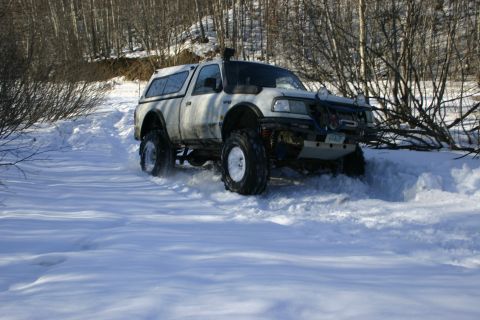 2003 Jeep TJ - A white pickup truck navigates a snow-covered path, its large tires kicking up snow as it drives through the winter landscape.