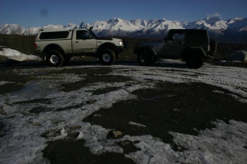 2003 Jeep TJ - Two modified off-road vehicles are parked on a dirt road with snow-capped mountains in the background.