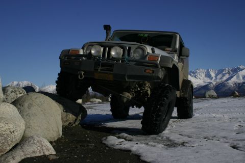2003 Jeep TJ - A silver Jeep TJ navigates rocky terrain with snow-covered mountains in the background.