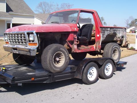 1978 Ford Bronco - A stripped red Ford body shell, doors, fenders and rear panels removed, one black bucket seat still in place, strapped to a black tandem-ax…