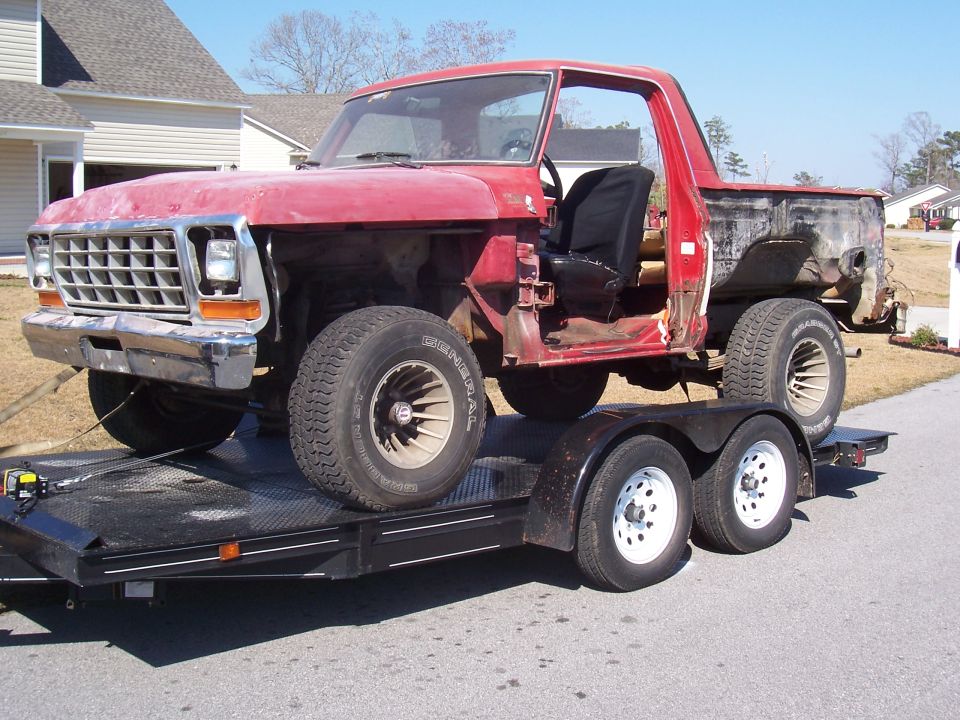 Stripped red Ford truck shell on a black tandem-axle trailer in a driveway