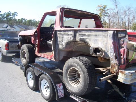1978 Ford Bronco - Three-quarter rear view of a gutted red full-size Bronco body shell strapped to a tandem-axle flatbed trailer in a suburban street, with no…