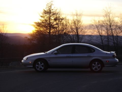 1999 Ford Taurus - A silver 1999 Ford Taurus sedan is parked on the side of a road as the sun sets behind distant hills and trees.