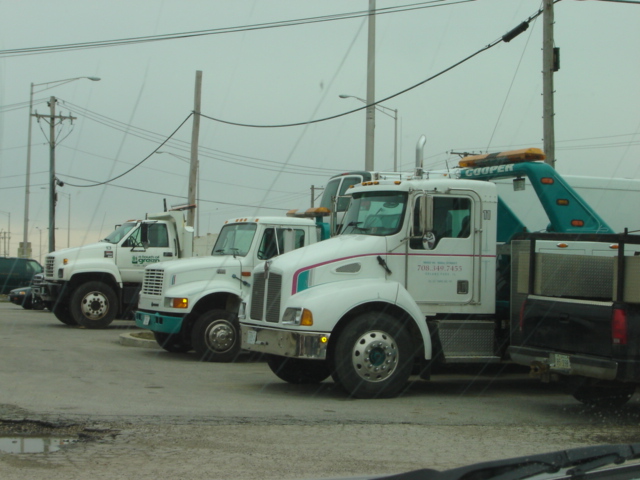 Three tow trucks parked side-by-side. The closest truck is white with red and blue stripes on the hood. A phone number is visible on the door