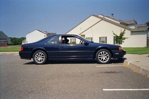 1989 Ford Thunderbird - Side profile of a dark blue 1989 Ford Thunderbird coupe parked in a lot with houses behind it.