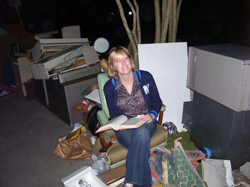 Woman reading a book while sitting in a chair amidst a pile of discarded appliances and junk