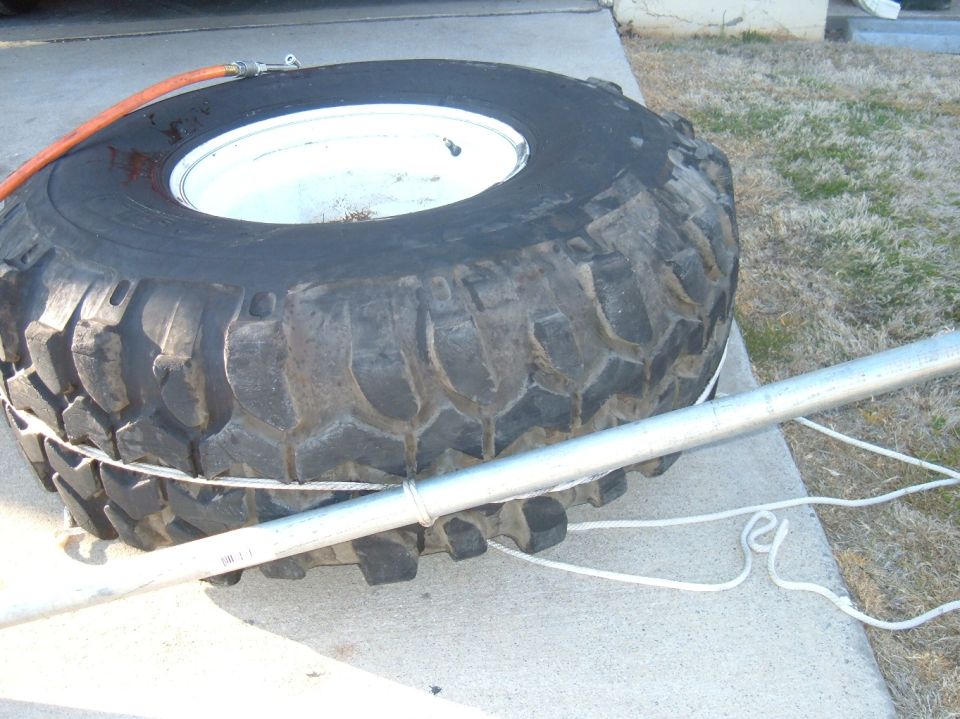 Large off-road tire with white rim, secured by a metal bar and rope