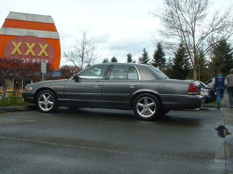 1999 Ford Crown Victoria - A gray Ford Crown Victoria is parked in front of a distinctive XXX Root Beer building.