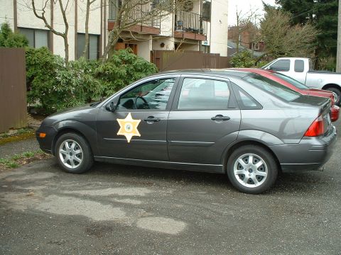 1999 Ford Crown Victoria - A dark gray sedan with a sheriff's star badge is parked next to a red car and a white pickup truck.