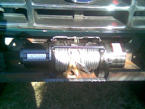 1992 Ford Bronco - Close-up of a black electric winch marked "REW10000" mounted on a steel plate behind the front bumper of a dark green Ford truck, with stee…