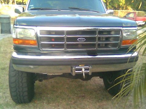 1992 Ford Bronco - Head-on view of a dark green 1992-96 Ford Bronco/F-series parked on grass, showing the chrome-trimmed grille with blue Ford oval, amber cor…