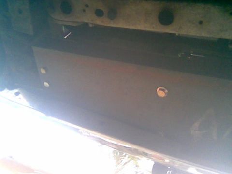 1992 Ford Bronco - Dim under-vehicle view of a flat steel mounting plate spanning behind the front bumper, showing bolt holes and a brass-colored fastener. Th…