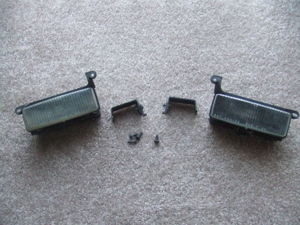 Two clear fog lights with black housings and mounting brackets, along with screws, on a carpeted surface
