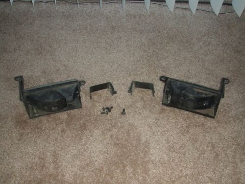 1995 Ford Probe - Two black plastic headlight housings, along with their mounting brackets and screws, are laid out on a carpeted surface.