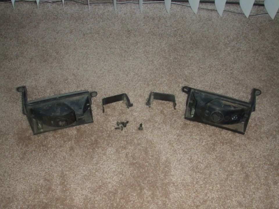 Two black plastic headlight housings with brackets and screws on carpet