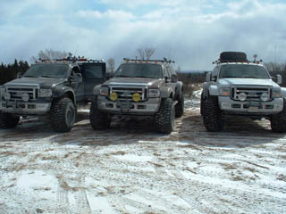 1984 Ford Bronco - Three pickup trucks are parked in a snowy outdoor area. The trucks appear to be modified for off-road use.
