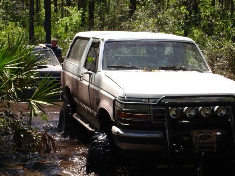 1989 Ford Bronco - A white Ford Bronco navigates through a muddy, wooded area with another vehicle partially visible behind it.