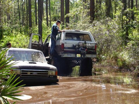 1989 Ford Bronco - Two off-road vehicles are stuck in a muddy forest clearing, with one Bronco partially submerged.