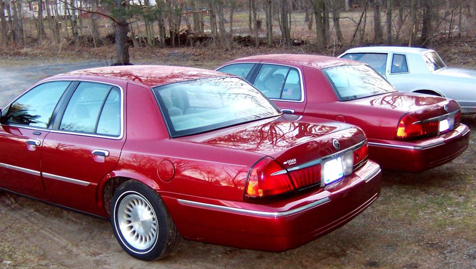 Two dark red Mercury Grand Marquis sedans parked side by side on gravel, viewed from the rear quarter