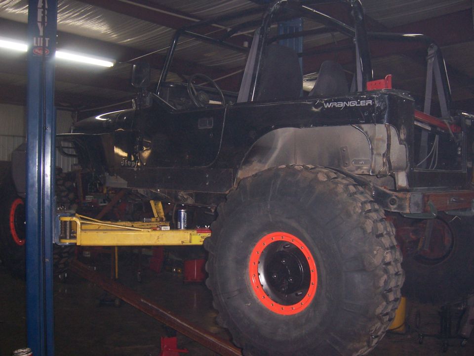 Black Jeep Wrangler mid-build in a shop, roll cage, cut fenders and mud tires on orange beadlock wheels