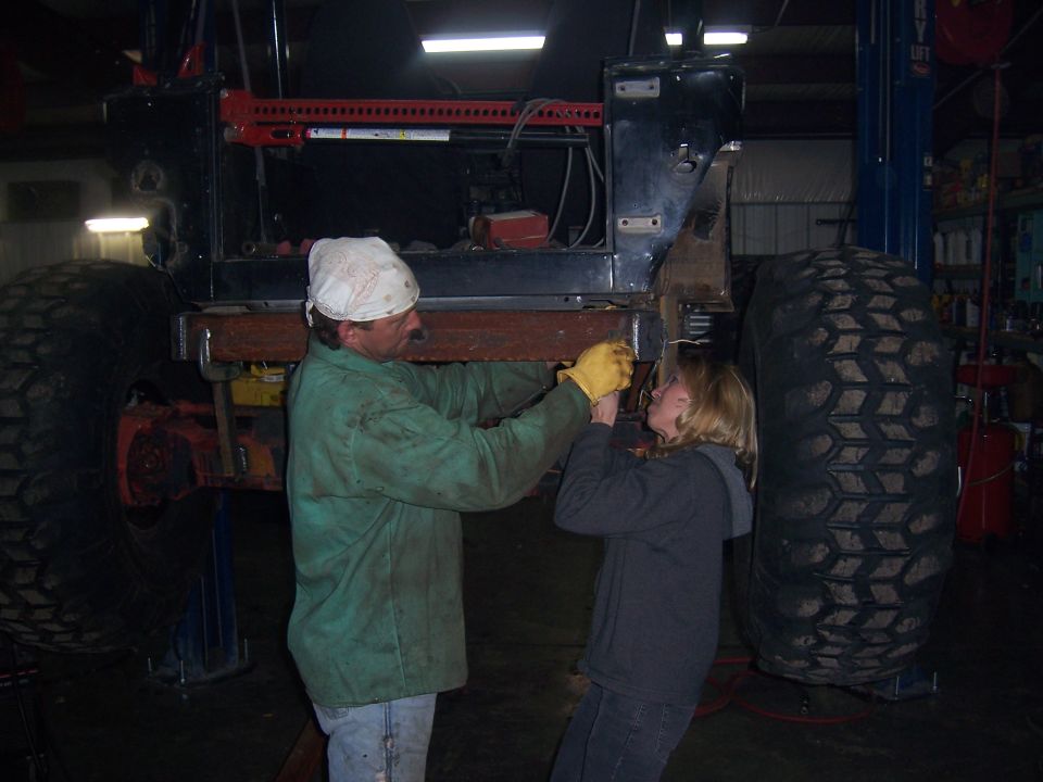 Two people fit a steel crossmember under a black Jeep tub raised on big mud tires in a shop