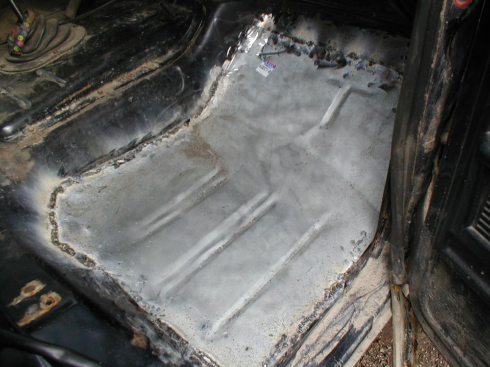 Interior floor pan of a vehicle undergoing repair, showing bare metal and welding marks. A small sticker is visible