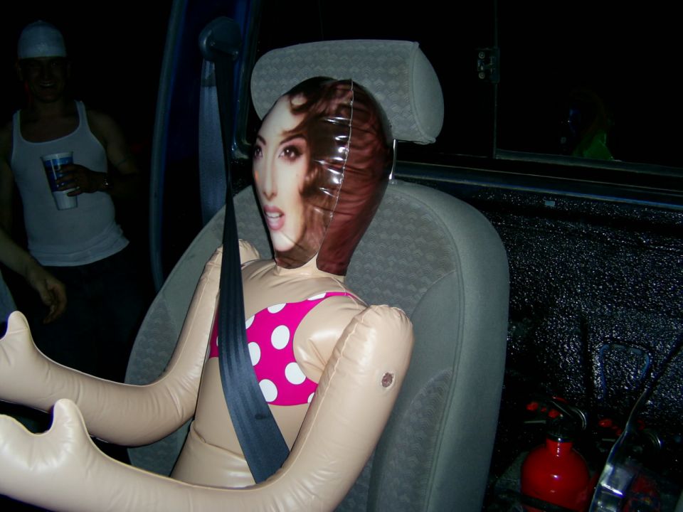 inflatable doll in car seat, wearing pink polka dot bikini, seatbelt on. guy in background holding cup