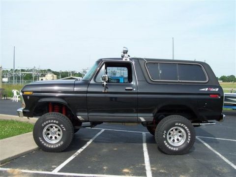 1978 Ford Bronco - Side profile of a lifted dark blue full-size Ford Bronco with removable hardtop, parked in an empty paved lot. It rides on Cooper A/T tires…