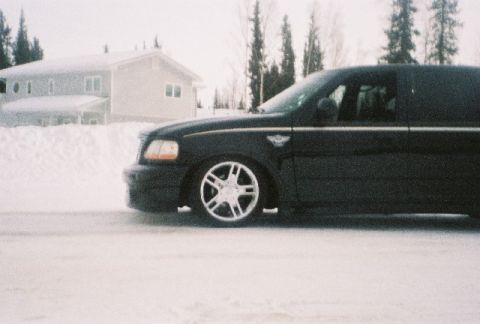 2003 Ford F-150 - A black 2003 Ford F-150 pickup truck is seen from the side, driving on a snowy road with a house in the background.