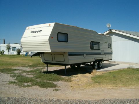 1985 Ford Bronco - A fifth wheel trailer with 'Sandpiper' branding is parked on a concrete pad next to a residential building.