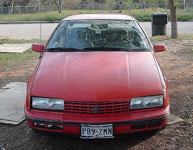 Red Chevrolet Beretta coupe, front view with Texas plate, by a chain-link fence