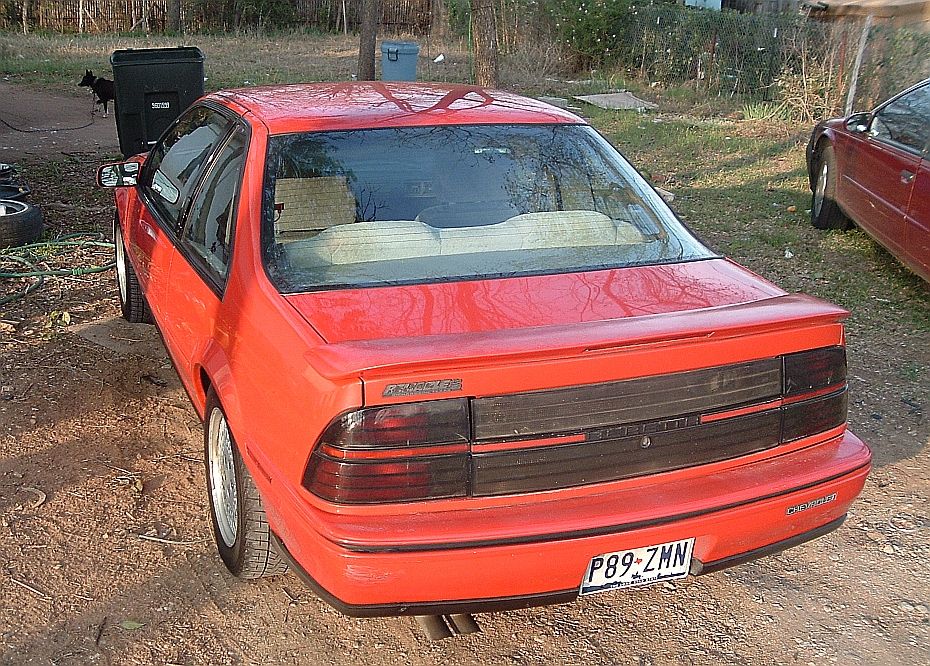 Red two-door coupe parked on dirt, rear three-quarter view with full-width tail lights