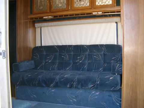 1985 Ford Bronco - The interior of a trailer features a blue sofa and wooden cabinets, with a window covered by a shade.