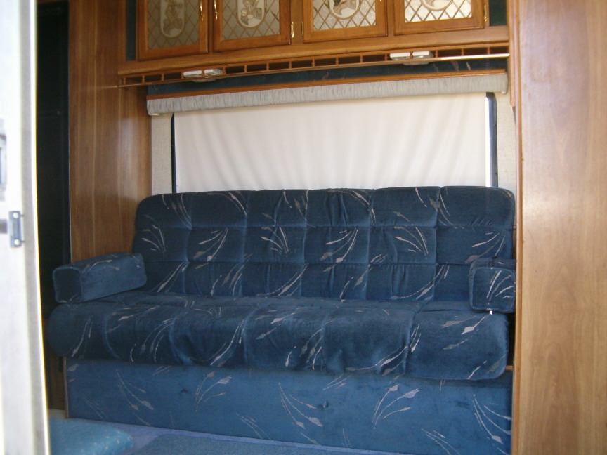 Interior of a trailer showing a blue sofa with a patterned fabric, wooden cabinets above, and a window with a pulled-down shade