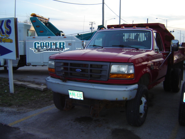 Red dump truck with a silver bumper and a gray grille. A tow truck is visible behind it