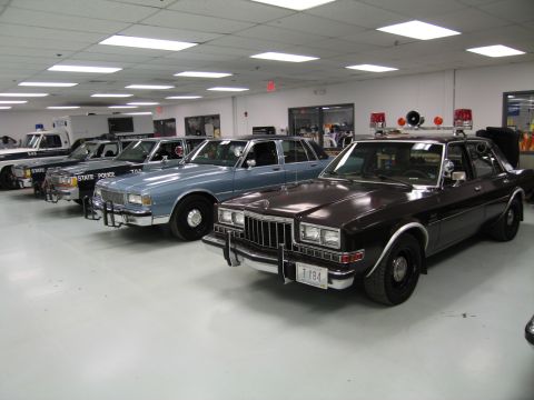 1991 Ford LTD Crown Victoria - Several vintage police sedans lined up inside a museum showroom, with a maroon-brown sedan wearing a light bar in the foreground.