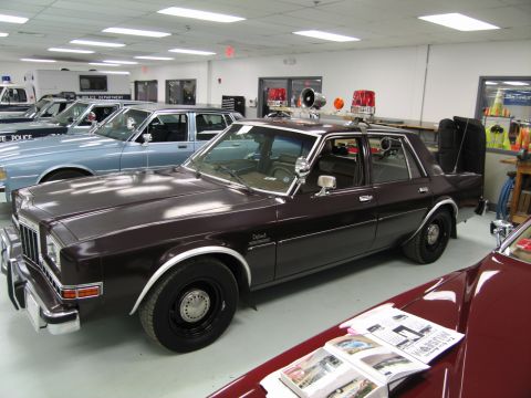 1991 Ford LTD Crown Victoria - brown four-door vintage police sedan with a roof light bar parked in a showroom, more cruisers visible behind it.