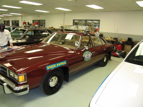 1991 Ford LTD Crown Victoria - maroon four-door cruiser marked 'STATE PATROL' with a door shield and roof light bar, displayed inside a showroom.