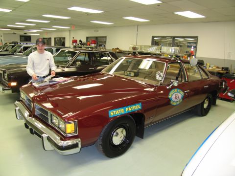 1991 Ford LTD Crown Victoria - man in a cap stands beside a maroon 'STATE PATROL' four-door sedan with a roof light bar in a showroom.
