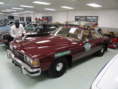 1991 Ford LTD Crown Victoria - person in a balaclava leans over the hood of a maroon 'STATE PATROL' sedan with a roof light bar inside a showroom.