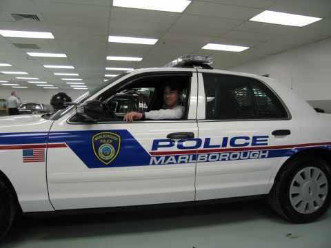 1991 Ford LTD Crown Victoria - man in a cap sits in the driver's seat of a white 'POLICE MARLBOROUGH' cruiser with light bar, viewed from the side.