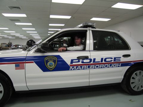 1991 Ford LTD Crown Victoria - person sits in the driver's seat of a white 'POLICE MARLBOROUGH' cruiser with roof light bar, shown in side profile.