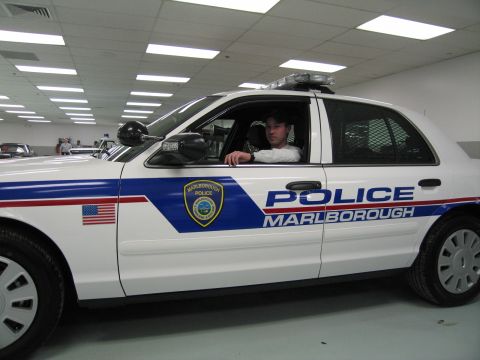 1991 Ford LTD Crown Victoria - man rests his arm out the window of a white 'POLICE MARLBOROUGH' cruiser with light bar, viewed from the side.