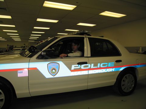 1991 Ford LTD Crown Victoria - person sits in a tan 'POLICE MARLBOROUGH' cruiser with red striping and roof light bar, photographed under warm indoor lighting.